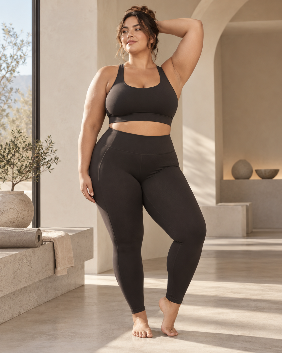Plus-size woman wearing espresso ROMY high-rise leggings and a black sports bra in a sunlit stone interior